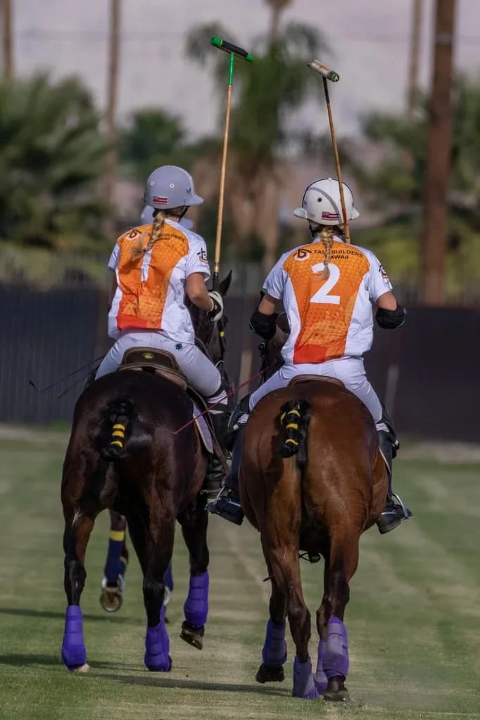 Two Apollo Team riders in Tall Builders Hawaiʻi jerseys, numbers 1 and 2, crossing the polo field